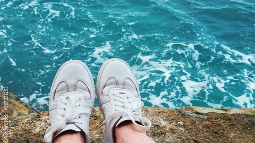 Traveler or adventurer, nomad urban explorer sits on cliff or rock in white sneakers, watches waves crush and roll over shore. Influencer or blogger lifestyle. Feet hanging from cliff waves on coast
