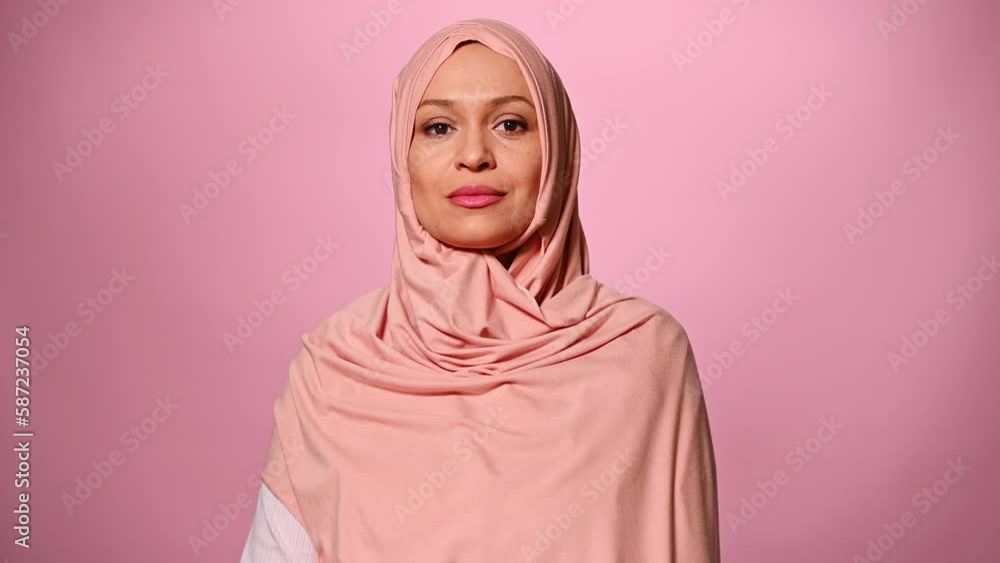 Closeup portrait of a confident beautiful middle-aged Arab Muslim woman in pink hijab, cutely smiling, looking at camera, isolated on pink background. Middle-Eastern female fashion. Islamic culture Closeup portrait of a confident beautiful middle-aged Arab Muslim woman in pink hijab, cutely smiling, looking at camera, isolated on pink background. Middle-Eastern female fashion. Islamic culture