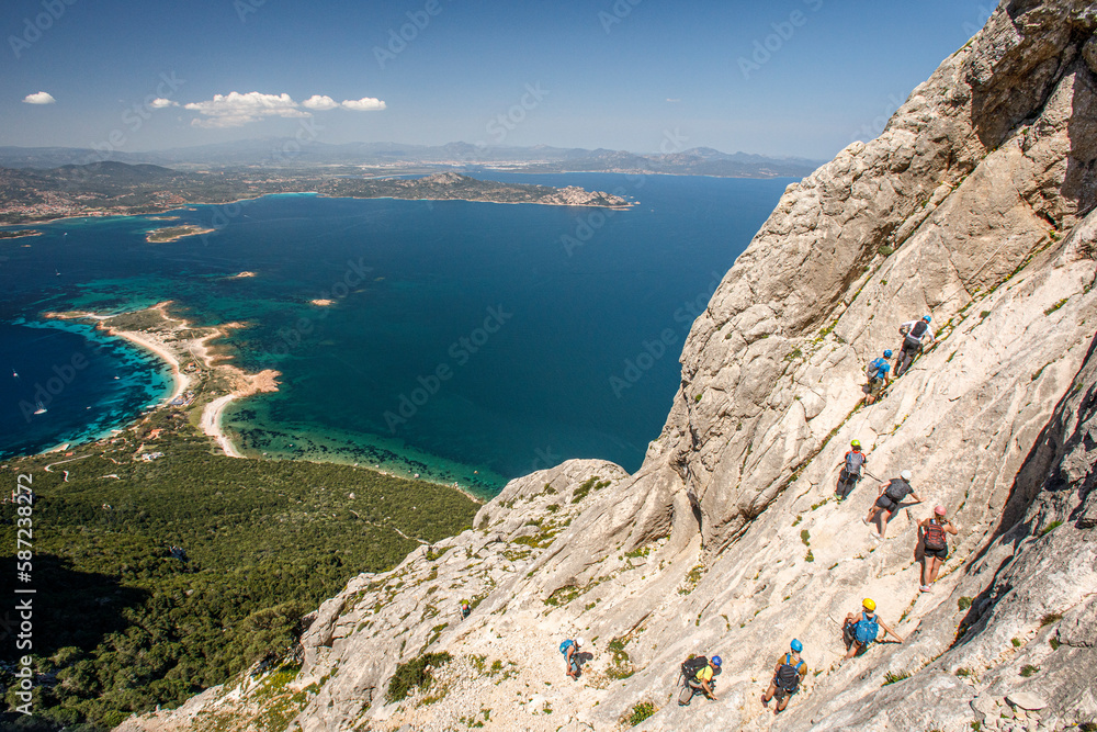 foto panoramica di una costa della Sardegna mentre un gruppo di escursionisti compie una ferrata nella parete rocciosa