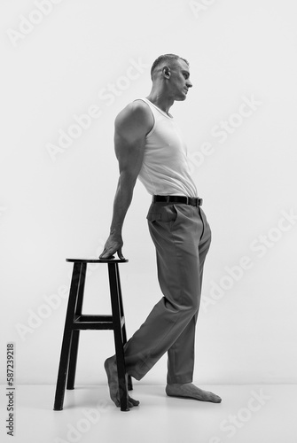 Portrait of elegant muscular man wearing pants and white t-shirt leaning on chair over studio background. Monochrome, side view