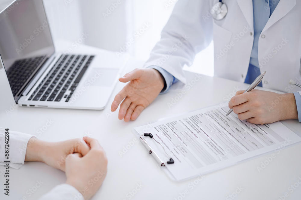 Doctor and patient sitting opposite each other at the desk in clinic. The focus is on female physician's hands filling up the medication history record form or checklist, close up. Medicine concept Doctor and patient sitting opposite each other at the desk in clinic. The focus is on female physician's hands filling up the medication history record form or checklist, close up. Medicine concept