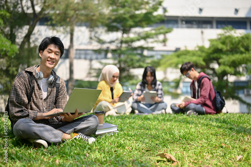 Smiling Asian male college student sits on the grass in the campus park with his laptop.