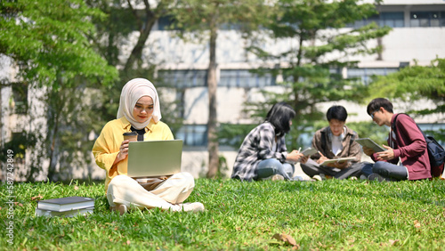 Focused Asian Muslim female college student in hijab sitting on the grass, using her laptop