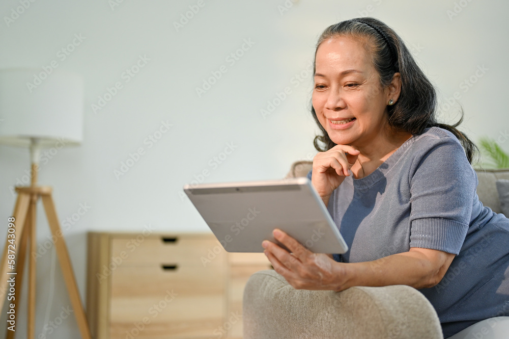 Happy Asian-aged retired woman watching movie on her digital tablet on sofa Happy Asian-aged retired woman watching movie on her digital tablet on sofa