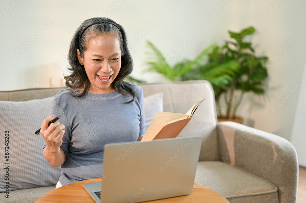 Naklejka premium Excited Asian-aged retired woman holding pen and book, using her laptop in living room