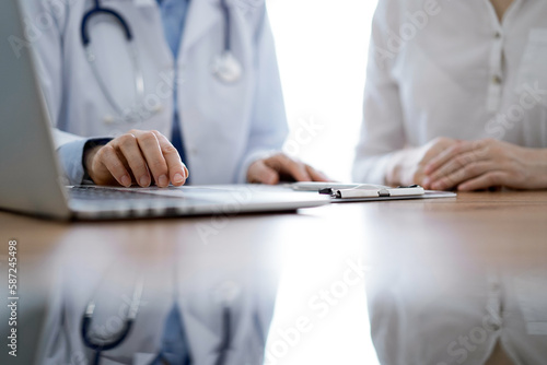 Doctor and patient discussing something while using laptop computer and sitting near each other at the wooden desk in clinic. Medicine concept