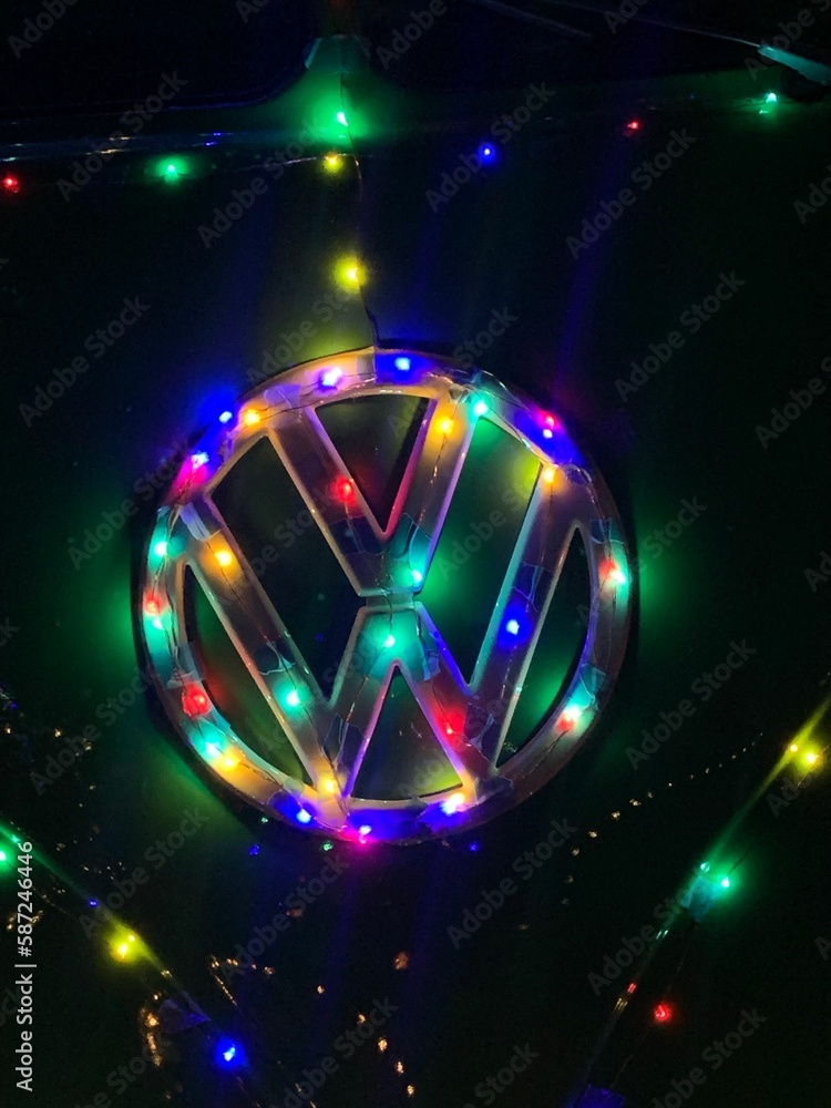 Obraz premium Vertical closeup of the Volkswagen logo on the vehicle surrounded with colorful Christmas lights