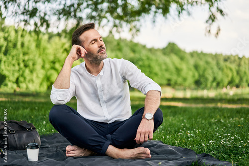 Middle-aged man sitting cross-legged on blanket in park daydreaming feeling relaxed