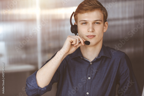 Young blond businessman using headset and computer in a darkened office, glare of light on the background. Startup business means working hard