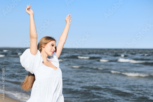 Happy smiling beautiful woman on the ocean beach standing in a white summer dress  raising hands