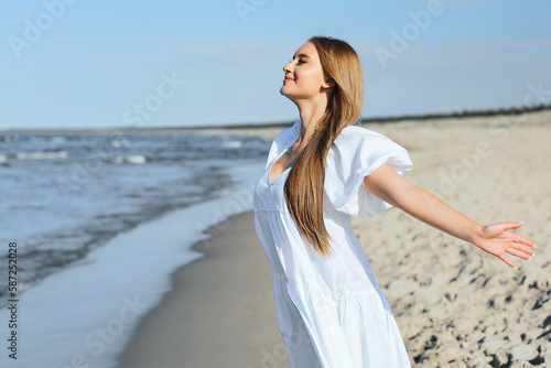 Happy smiling beautiful woman is on the ocean beach in a white summer dress, open arms