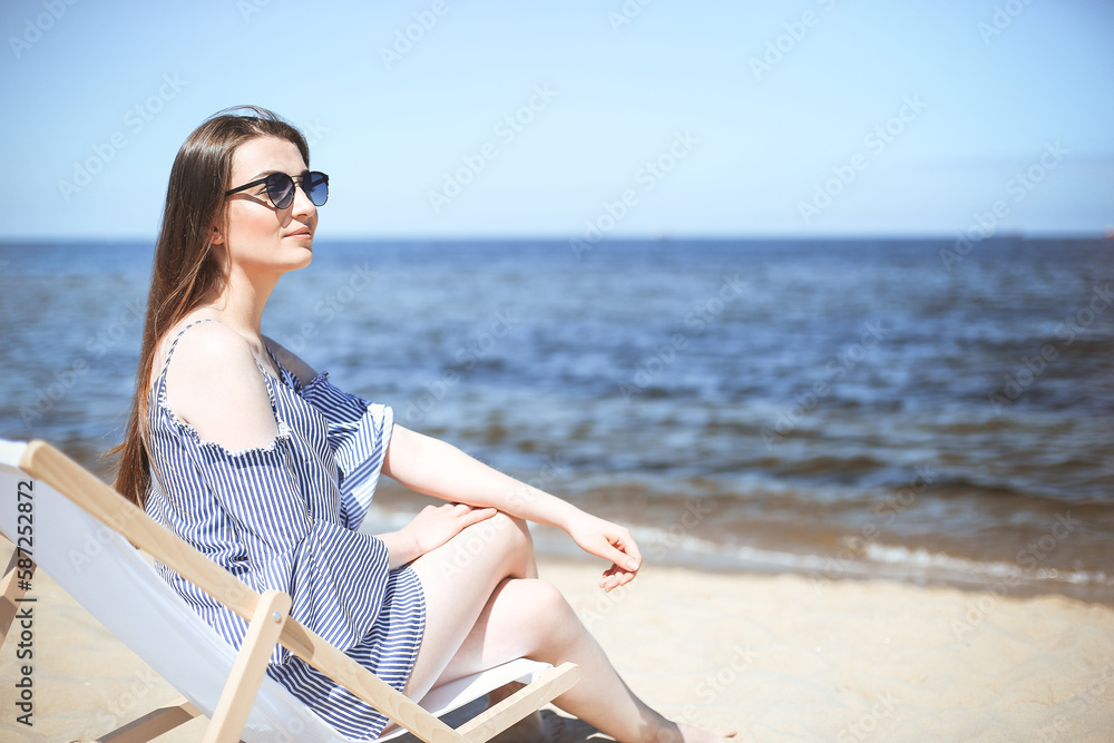 Happy young brunette woman relaxing on a wooden deck chair at the ocean beach while smiling, and wearing fashion sunglasses. The enjoying vacation concept