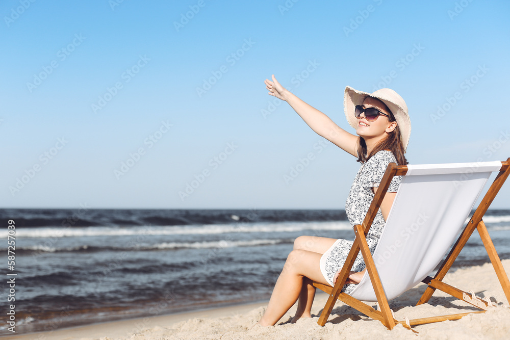 Happy brunette woman sitting on a wooden deck chair at the ocean beach while waving and greeting somebody with her hand