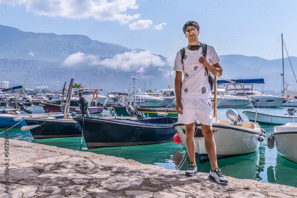 Young adult male tourist posing near the boats moored on the backdrop of the Dukley Marina port in Budva, Montenegro. Man in white shorts and a T-shirt stands on the backdrop of the seascape