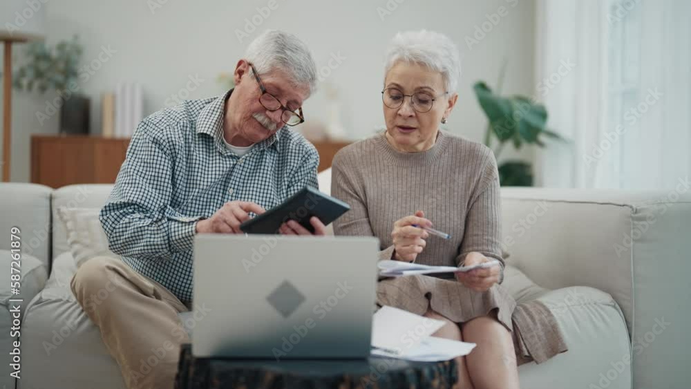 Senior couple calculating bills sitting on couch at home doing online payments on laptop. Do paperwork discuss unpaid taxes, overdue payment household expenses. Financial difficulties money problem.