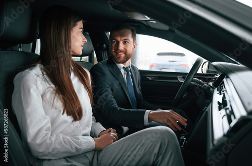 Man in suit and tie is sitting with young woman, customer, in the automobile and showing the interior of the car © standret