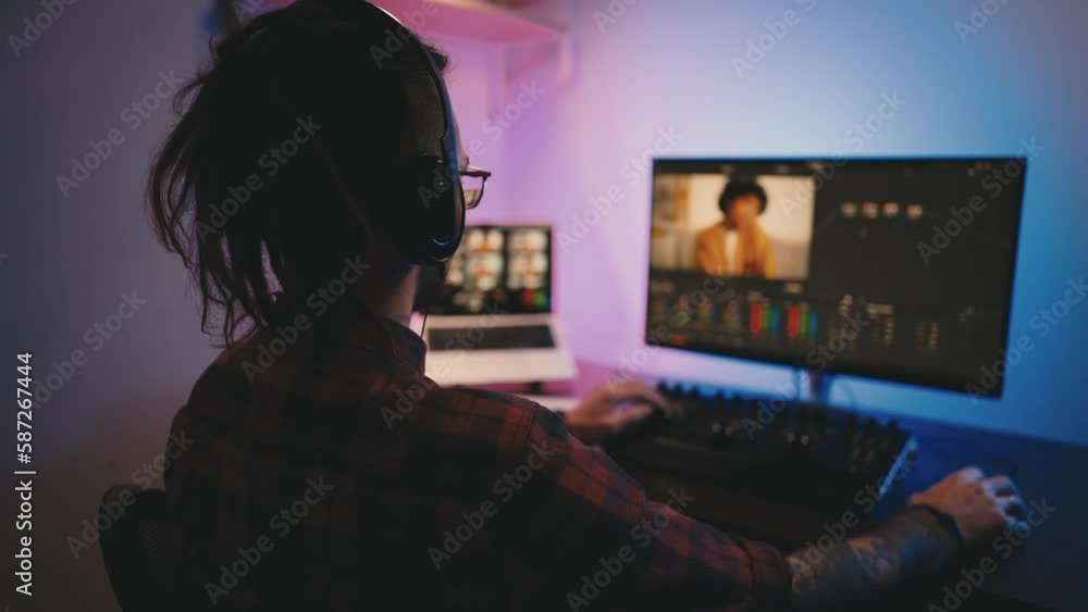 Man video editor wearing headphones sits at computer with backlight, looks in monitor. Video editing with music and color correction. Freelance, working in home office . Professional video processing. Man video editor wearing headphones sits at computer with backlight, looks in monitor. Video editing with music and color correction. Freelance, working in home office . Professional video processing.