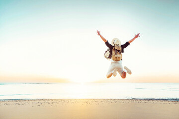 Happy traveler enjoying freedom jumping at the beach - Cheerful hiker with backpack raising hands up at sunset - Wellbeing, happiness, summer holidays and travel concept