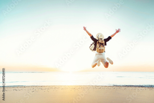 Happy traveler enjoying freedom jumping at the beach - Cheerful hiker with backpack raising hands up at sunset - Wellbeing, happiness, summer holidays and travel concept