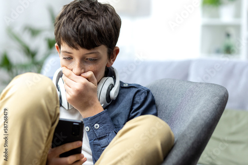 Young teenager boy reading bad news on phone, son sitting on sofa in living room with headphones at home closeup.