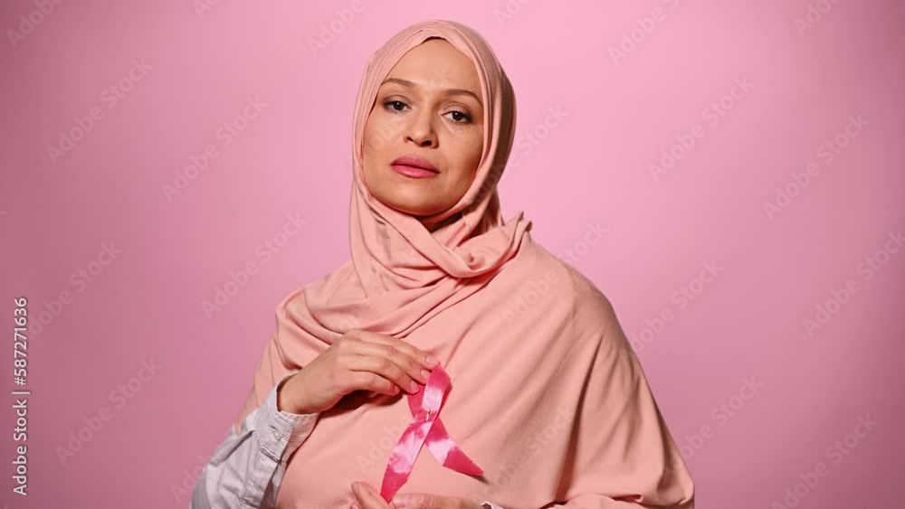Muslim woman in hijab, putting on pink satin ribbon, symbol of World Cancer Awareness Day