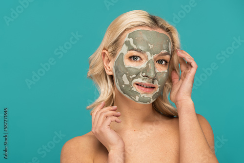 blonde woman with clay mask on face looking at camera isolated on turquoise.
