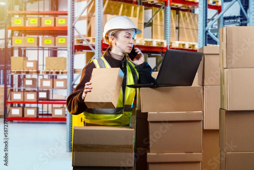 Warehouse of courier company. Woman works in delivery service warehouse. Girl prepares goods for shipment. Courier warehouse worker talking on phone. Female holding parcel and looking at laptop