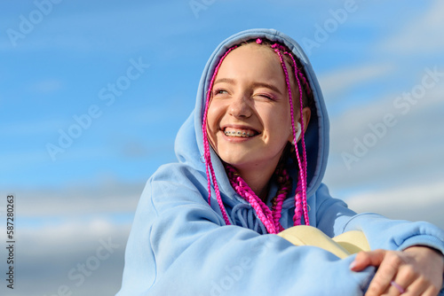 Portrait of a caucasian teenage girl with pink braids using wireless headphones against the blue sky.Technology,summer concept.Generation Z style. © Tatyana