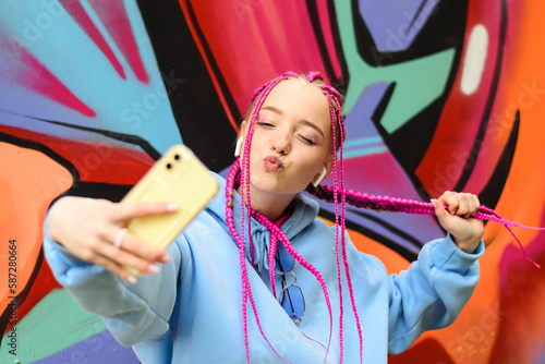 Caucasian teenage hipster girl with pink braids is using a smartphone against the background of a multicolored street wall.Summer concept.Generation Z style.Social media concept. photo