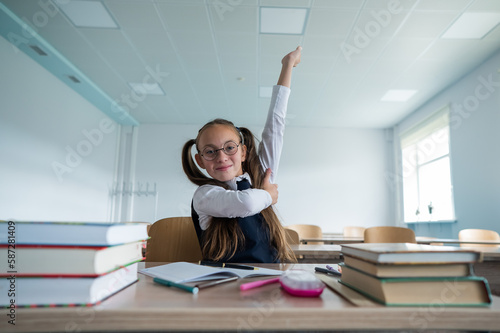 Caucasian girl with two ponytails sits at her desk and raises her hand up to answer the teacher's question.