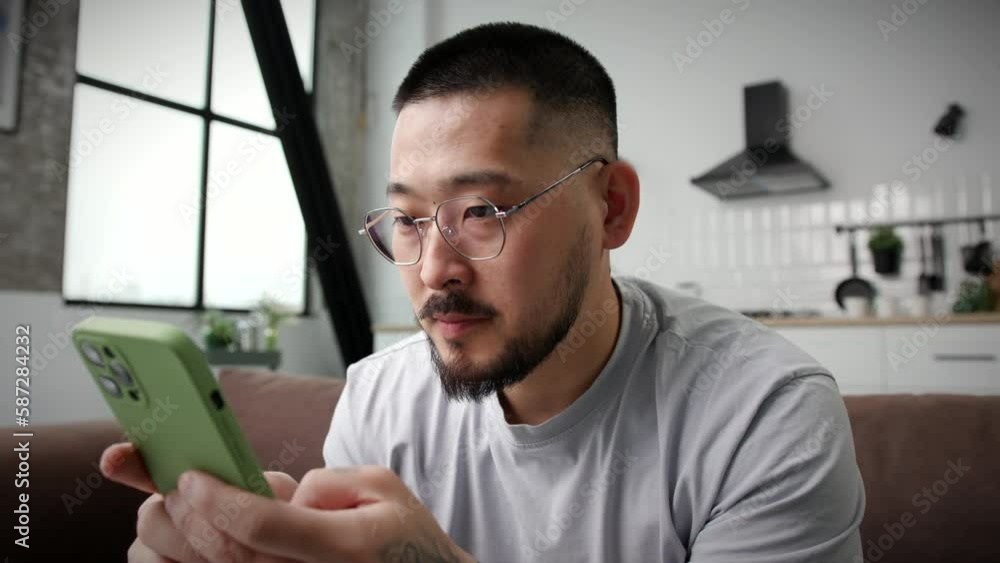 Smiling Man Texting Using Smartphone At Home Close Up. Korean man chatting online holding cell phone in hands sitting on couch. Smiling Man Texting Using Smartphone At Home Close Up. Korean man chatting online holding cell phone in hands sitting on couch.