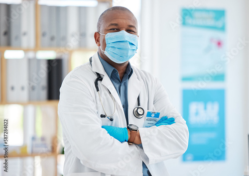 Covid  portrait and a doctor black man in a hospital office  standing arms crossed for healthcare or insurance. Medical  mask and trust with a male medicine professional at work in a wellness clinic