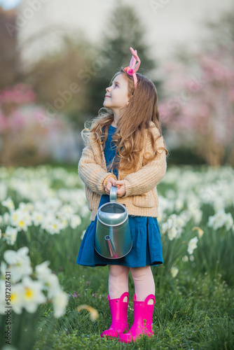 Easter. Cute little girl of 5 years old with rabbit ears is watering daffodils on the lawn. Happy child. Spring holidays.