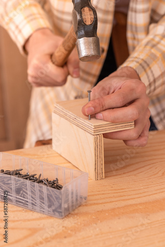 Handyman hammering nail into wooden plank. Crop anonymous male woodworker using hammer to drive nail into wooden board on workbench near box with nails