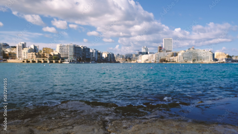 Blurred City buildings with blue sea water and blue sky with clouds. Blur view of the city with blue sea at sunny day time. 