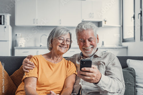 Happy mature family, wife and husband using phone together at home, smiling middle aged man and woman sitting on couch, using mobile device apps, watching video in social network, surfing internet.