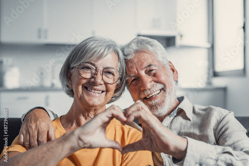 Close up portrait happy sincere middle aged elderly retired family couple making heart gesture with fingers, showing love or demonstrating sincere feelings together indoors, looking at camera..