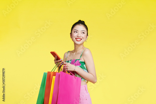 Young excited cheerful woman holding shopping bags isolated over yellow background using mobile phone looking aside.