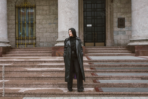 Street photography. A beautiful stylish girl dressed in all black, wearing a long leather coat or jacket, stands in front of a large building with columns.