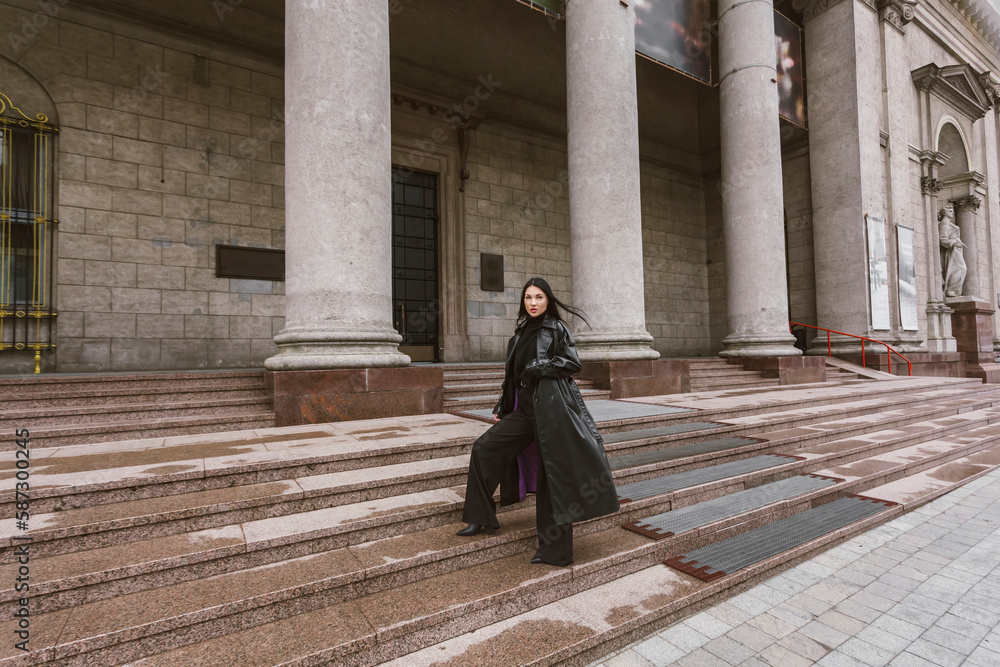 Street photography. A beautiful stylish girl dressed in all black, wearing a long leather coat or jacket, is climbing the steps of a large building with columns.