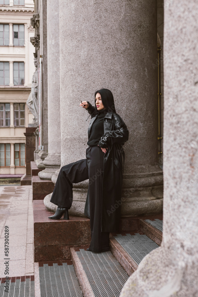 Street photography. A beautiful stylish girl dressed in all black, wearing a long leather coat or jacket, is posing against of large building columns.