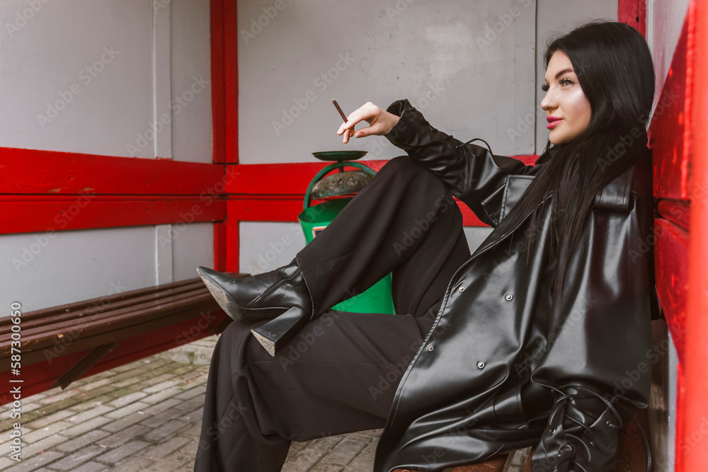 Naklejka premium Street photography. A beautiful girl dressed in all black, wearing a long leather coat or jacket, sits on an old dirty bus stop with a cigarette in her hand.