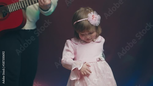 Little girl on stage in pink vintage dress, she dances, her father plays acoustic guitar. Color music is shining. Performance on stage. Early development of children