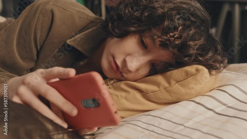 Loose close-up of sad young girl with curly short dark hair lying in bed, head on yellow pillow, checking her phone, then putting it aside looking depressed.