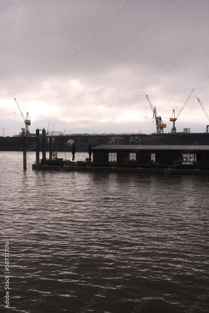 Vertical monochrome of a floating cafe on the water before the cranes in the port of Hamburg
