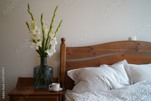 Bedroom closeup view. Spring blooming branch, white flowers on the bedside table, cup of tea or coffee and white bedding sheets. Spring, light, calmness, coziness.
