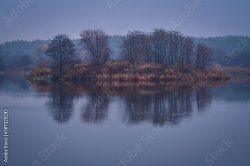 Morning foggy landscape over the water. Trees on an islet on a cloudy day at Lake Mojcza in Kielce, Poland.