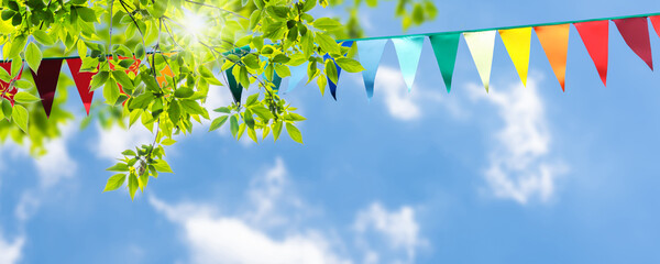 colorful pennant string decoration in green tree foliage on blue sky, summer party background template banner with copy space