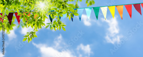 colorful pennant string decoration in green tree foliage on blue sky, summer party background template banner with copy space