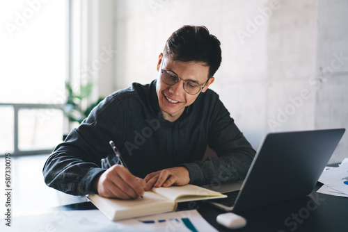 Happy young man writing in notepad on table with laptop